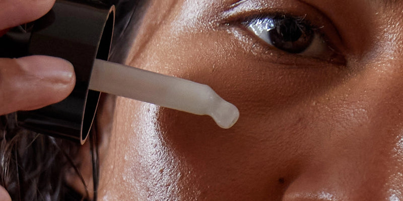 Nahaufnahme einer Person, die ein Hautpflegeserum mit einer Pipette auf ihre Wange aufträgt, wobei der Schwerpunkt auf den Augen und der glatten Hautstruktur liegt.