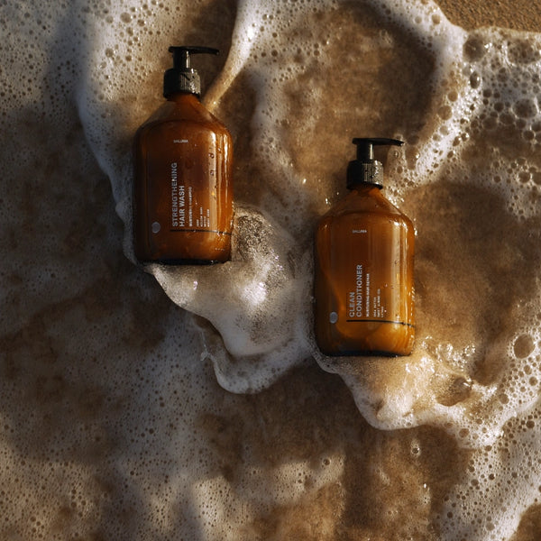 Zwei braune Pumpflaschen mit weißen Etiketten liegen auf dem nassen Sand, während Meeresschaum über sie hinwegspült. Die Flaschen scheinen Körperpflegeprodukte wie Shampoo oder Seife zu sein.