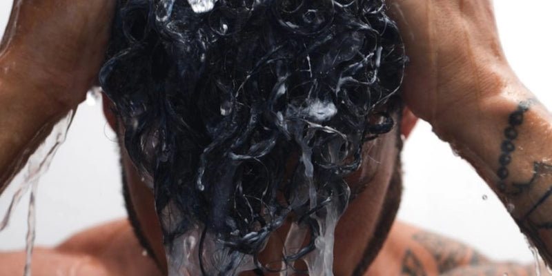 Eine Person mit lockigem, dunklem Haar wäscht sich die Haare, wobei das Wasser herunterläuft, während sie mit beiden Händen ihre Kopfhaut massiert. Auf ihrem linken Arm sind Tätowierungen zu sehen.
