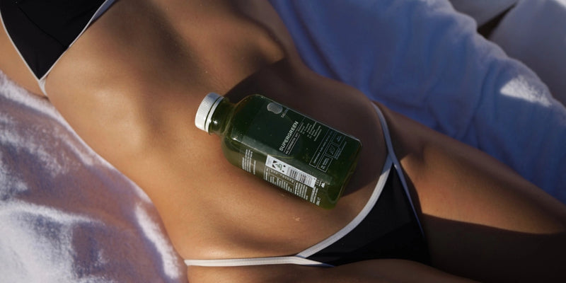 Eine Person, die einen schwarzen Bikini trägt, liegt auf einem weißen Handtuch und hat eine grüne Saftflasche auf dem Bauch liegen, die von der Sonne angestrahlt wird.