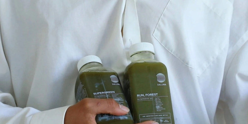 Eine Person mit weißem Hemd hält zwei grüne Saftflaschen mit den Aufschriften SUPERGREEN und RUN, FOREST und weißen Kappen.