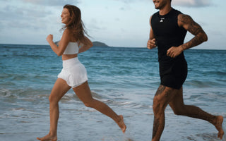Lachende Frau in weißem Sportoutfit und Mann in schwarzem Sportoutfit joggen barfuß am Strand durch die Brandung, im Hintergrund Meer und eine kleine Insel