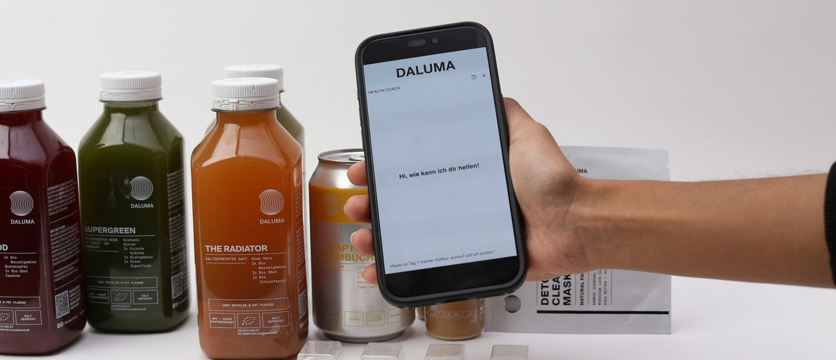 Smartphone mit dem DALUMA Health Coach Chat vor kaltgepressten Säften, Kombucha und Detox Clearing Mask.