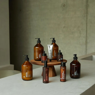 Eine Reihe von bernsteinfarbenen und braunen Hautpflegeflaschen und -tuben, die auf einem kleinen Holzständer vor einem minimalistischen Betonhintergrund angeordnet sind.
