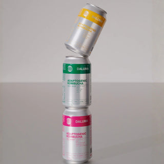 Drei Dosen Daluma Adaptogenic Kombucha sind auf einer spiegelnden Oberfläche gestapelt, mit gelben, grünen und rosafarbenen Etiketten, und die oberste Dose ist schräg gestellt.