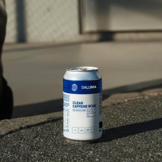 Eine Dose Daluma Clean Caffeine BCAA-Energydrink steht auf einer rauen Außenfläche im Sonnenlicht, mit einem Zaun und Beton im unscharfen Hintergrund.