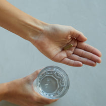 Eine Person hält eine durchsichtige Kapsel von DALUMA 360 DAILY ESSENTIALS SUPPLEMENT in einer Hand und ein Glas Wasser in der anderen, vor einem schlichten hellen Hintergrund, der die natürlichen Inhaltsstoffe hervorhebt.