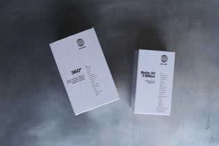 Zwei weiße rechteckige DALUMA-Ergänzungsdosen, darunter 360 DAILY ESSENTIALS SUPPLEMENT, ruhen auf einer Metalloberfläche. Beide haben ein minimalistisches Design, ein rundes Logo, Inhaltsstofflisten und heben natürliche Inhaltsstoffe hervor.