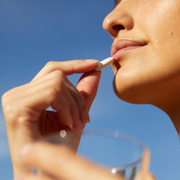 Nahaufnahme von Gesicht und Händen einer Person, die eine FACE GLOW DUO-Kapsel von DALUMA an die Lippen hält, mit einem Glas Wasser in der anderen Hand und einem klaren blauen Himmel im Hintergrund.