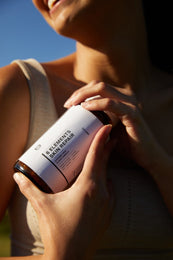 Eine Frau in einem beigen Tank-Top hält einen Tiegel mit der Aufschrift FACE GLOW DUO von DALUMA an ihre Brust und lächelt sanft in das natürliche Sonnenlicht vor einem blauen Himmel, um die Vorteile dieser Hautpflege hervorzuheben.