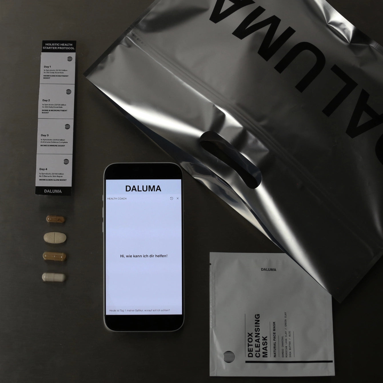 Ein Foto zeigt eine Tasche der Marke DALUMA, einen Ergänzungsstreifen mit vier Kapseln, ein Smartphone mit der DALUMA-Website und einen silbernen Beutel mit der Aufschrift 
