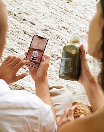 Zwei Personen sitzen auf einem Teppich, eine hält ein Smartphone mit der Galuma-App in der Hand, die andere hält eine durchsichtige Flasche mit einem grünen Saft oder Smoothie in der Hand.