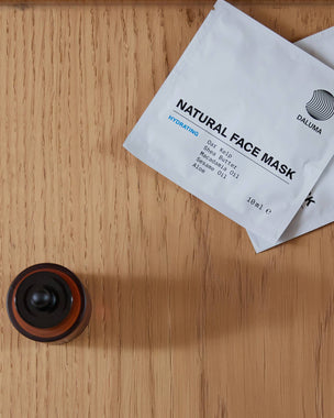 Eine Flasche mit einer Pipette und zwei weiße Päckchen mit der Aufschrift Natural Face Mask Hydrating stehen auf einer Holzfläche. Der Markenname Daluma ist auf den Packungen sichtbar.