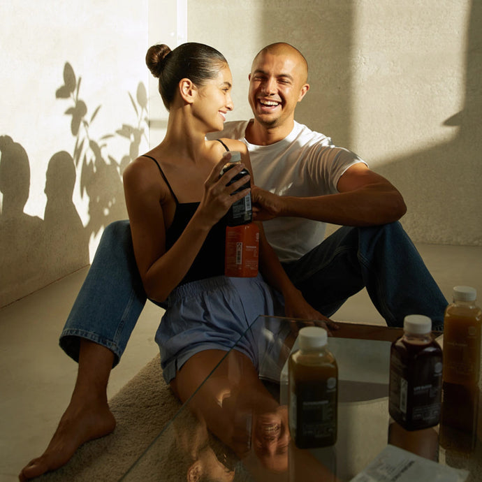 Eine Frau und ein Mann sitzen auf dem Boden, lächeln und lachen zusammen mit Getränken in Flaschen. Das Sonnenlicht fällt ein und wirft weiche Schatten. Vor ihnen auf dem Glastisch stehen mehrere Flaschen des DALUMA DAILY FEEL GOOD JUICE PACKAGE.