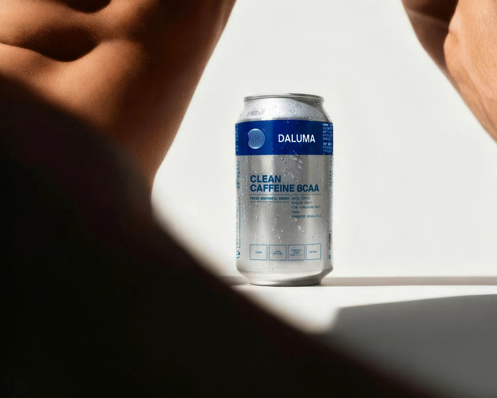 Eine Dose DALUMA CLEAN BCAA ENERGY DRINK aus dem 24er-Pack steht auf einer weißen Fläche, die Tropfen glitzern, eingerahmt von den verschwommenen Silhouetten zweier durchtrainierter, nackter Oberkörper.