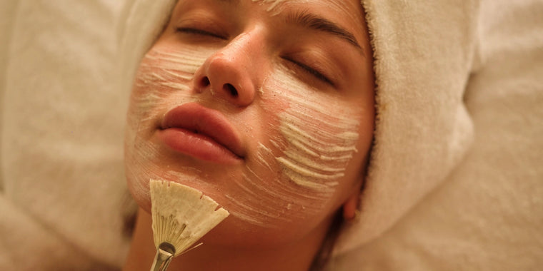 Eine Person liegt mit einem Handtuch um den Kopf gewickelt, die Augen geschlossen, während eine Gesichtsmaske mit einem Pinsel auf das Gesicht aufgetragen wird.
