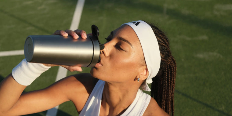 Eine Frau, die ein weißes Stirnband und ein Armband trägt, trinkt aus einer Edelstahlflasche, während sie auf einem Tennisplatz im Sonnenlicht sitzt.