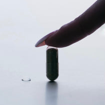 Eine Hand mit polierten Nägeln drückt sanft auf eine DALUMA IMMUN DEFENCE COMPLETE KOMPLEX DOUBLE Kapsel - reich an Kalmeghblattextrakt - die auf einer hellen Fläche neben einem Wassertropfen steht, um das Immunsystem zu stärken.