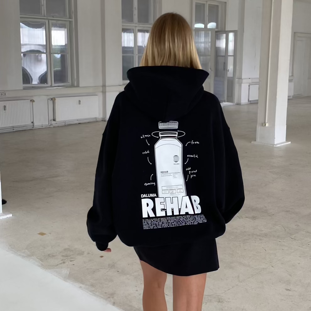 Eine blonde Person geht im Haus spazieren und trägt den DALUMA JOSEPH ATELIER X DALUMA REHAB HOODIE mit einer auffälligen Rückengrafik und dem Schriftzug 