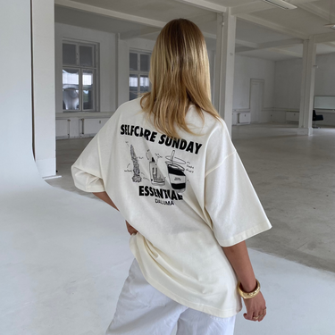 Eine Person mit langen blonden Haaren steht in einem minimalistischen weißen Raum und trägt das DALUMA JOSEPH ATELIER X DALUMA SELFCARE SUNDAY TEE - ein cremefarbenes Hemd aus Biobaumwolle in Übergröße mit der Aufschrift SELFCARE SUNDAY ESSENTIALS" auf dem Rücken - und dazu eine weiße Hose.