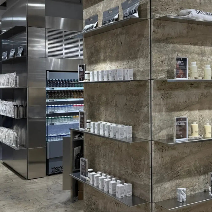 Modernes Einzelhandelsgeschäft mit Glasregalen, in denen Kosmetika und Hautpflegeprodukte übersichtlich angeordnet sind. Im Hintergrund ist ein Kühlregal mit Flaschenware zu sehen, daneben Metall- und Steinoberflächen.