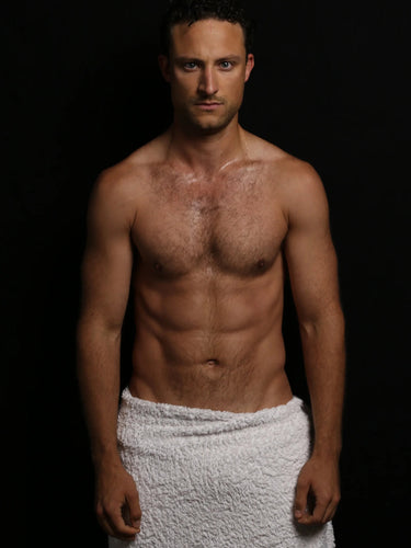 Ein Mann ohne Hemd mit kurzem, lockigem Haar steht vor einem schwarzen Hintergrund und blickt direkt in die Kamera. Er hat ein weißes Handtuch um seine Taille gewickelt.