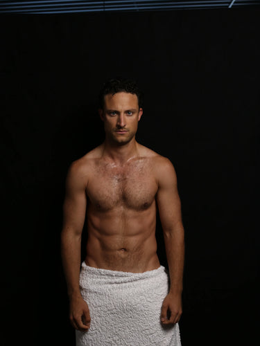 Ein Mann ohne Hemd und mit kurzen dunklen Haaren steht vor einem schwarzen Hintergrund und trägt nur ein weißes Handtuch um die Taille gewickelt. Er blickt mit ernster Miene nach vorne. Am oberen Rand des Bildes sind blaue horizontale Linien zu sehen.