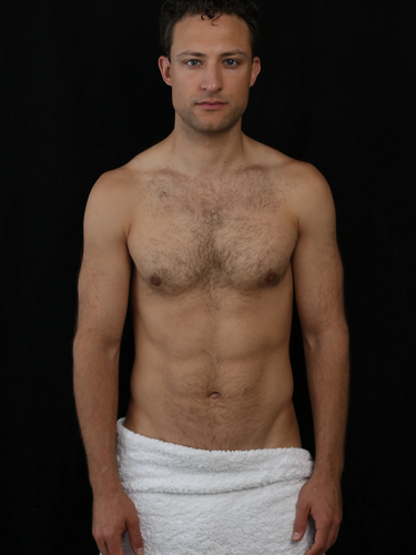 Ein Mann ohne Hemd und mit kurzen lockigen Haaren steht vor einem schwarzen Hintergrund und trägt ein weißes Handtuch um die Taille gewickelt. Er blickt mit neutraler Miene nach vorne.