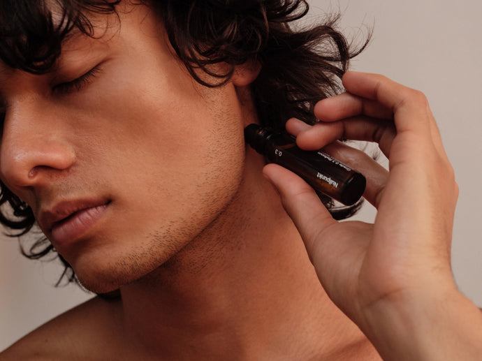 Eine Person mit mittellangem, lockigem Haar trägt Öl aus einem kleinen dunklen Fläschchen seitlich auf ihren Nacken auf, die Augen geschlossen und mit einem ruhigen, entspannten Ausdruck.