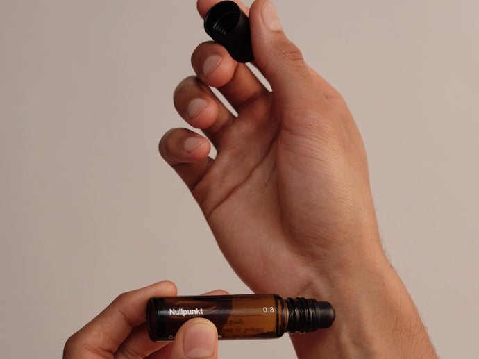 Eine Hand hält eine geöffnete bernsteinfarbene Rollerflasche mit der Aufschrift "Nullpunkt", während die andere Hand den Deckel abnimmt, vor einem schlichten Hintergrund.