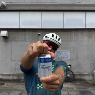 Ein Radfahrer mit Helm und Sonnenbrille hält eine Dose DALUMA CLEAN BCAA ENERGY DRINK 24 PACK mit grünem Kaffee und veganen BCAAs in die Kamera und verdeckt dabei teilweise sein Gesicht. Im Hintergrund sind ein Fahrrad und eine graue Betonwand zu sehen.