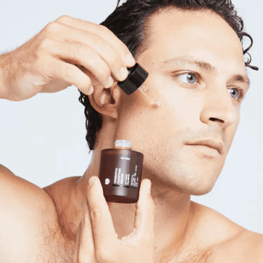 Ein Mann mit lockigem dunklem Haar trägt mit einer Pipette DALUMA PREBIOTIC PURIFYING GESICHTSSERUM aus einer braunen Flasche auf seine Wange auf, um seine Hautbarriere zu unterstützen, vor einem hellen Hintergrund.