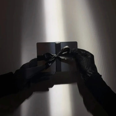 Eine Person mit schwarzen Handschuhen hält eine kleine schwarze Geschenkschachtel mit einer schwarzen Schleife - möglicherweise enthält sie den DALUMA THE GOOD VOUCHER GUTSCHEIN (30€, 50€ oder 100€) -, die von einem schmalen Lichtstrahl vor einem dunklen Hintergrund beleuchtet wird.