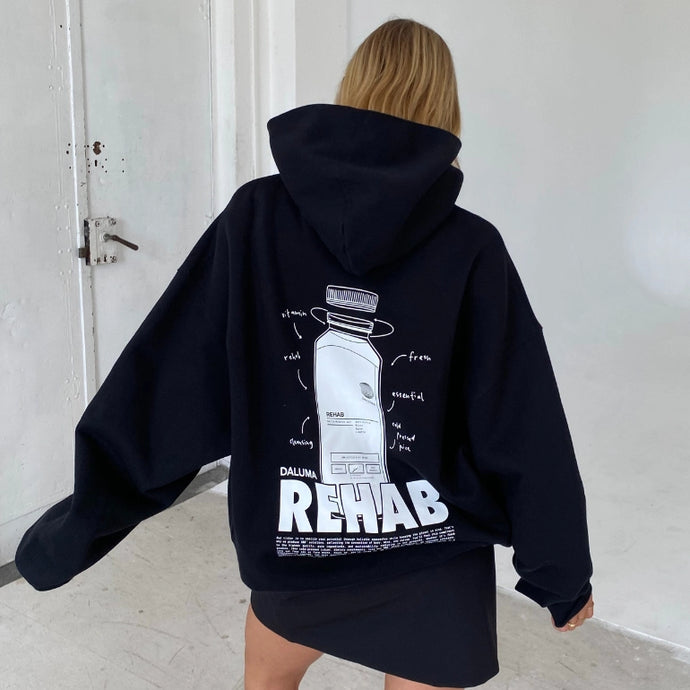 Eine Person mit langen blonden Haaren, die einen schwarzen Kapuzenpulli mit einem großen grafischen Muster und dem Wort REHAB auf dem Rücken trägt, steht drinnen in einem minimalistischen weißen Raum und blickt von der Kamera weg.