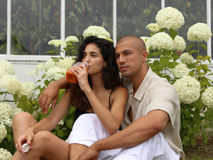 Eine Frau trinkt aus einer Flasche, während sie neben einem Mann sitzt, der seinen Arm um sie gelegt hat. Sie sitzen auf dem Boden vor weißen Hortensienblüten und einem Gewächshaus, beide wirken ruhig und entspannt.