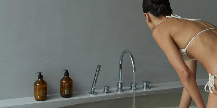 Eine Frau in einem weißen Bikini lehnt sich über den Rand einer Badewanne mit fließendem Wasser. Zwei braune Pumpflaschen stehen auf dem Badewannenrand neben ihr vor einer schlichten grauen Wand.