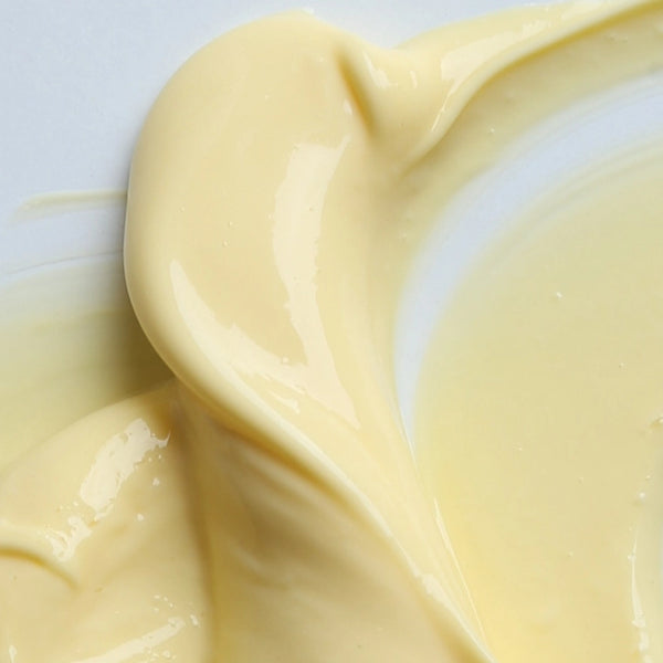Nahaufnahme einer glatten, cremigen, blassgelben Substanz, die sich auf einer weißen Oberfläche ausbreitet und an Vanillepudding, Sahne oder eine dicke Soße mit glänzender Textur erinnert.