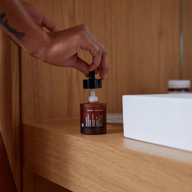 Eine Hand mit einer sichtbaren Tätowierung entnimmt mit einer Pipette die ANTI AGING ESSENTIAL ROUTINE von DALUMA aus einer braunen Glasflasche, die auf einer hölzernen Oberfläche neben einem weißen, rechteckigen Gegenstand steht und diesen wichtigen Anti-Aging-Schritt hervorhebt.