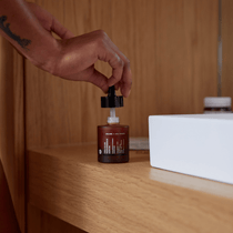 Eine Hand hält einen Tropfer über einer bernsteinfarbenen Flasche DALUMA ANTI AGING ESSENTIAL ROUTINE mit dunklem Etikett, die auf Holz neben einem weißen Behälter steht. Den Hintergrund bildet eine Holztäfelung.