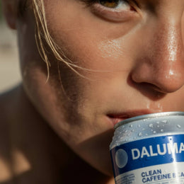 Nahaufnahme einer hellhaarigen, hellhäutigen Person, die eine Dose DALUMA CLEAN BCAA ENERGY DRINK 24 PACK an die Lippen hält, wobei das Sonnenlicht ihr Gesicht hervorhebt, während Kondenswasser auf der Dose glitzert.