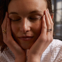 Eine Frau mit geschlossenen Augen und ruhigem Gesichtsausdruck stützt ihr Gesicht in ihre Hände, in denen sie goldene und silberne Ringe trägt. Ihr ruhiger Blick spiegelt die beruhigende Wirkung des CELLULAR RESURGENCE ANTI AGING SERUM von DALUMA auf die Hauterneuerung wider. Sie trägt ein weißes, strukturiertes Oberteil.