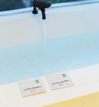 Aus einem schwarzen Wasserhahn fließt Wasser in eine Badewanne. Davor liegen zwei Packungen DALUMA CLEANSING AKTIVKOHLE FACE MASK auf dem weißen Wannenrand. Durch das Fenster hinter dieser entspannenden Szene sind draußen Bäume zu sehen.