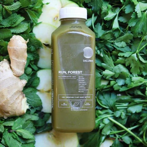 Eine Flasche des DAILY FEEL GOOD JUICE PACKAGE von DALUMA wird zwischen Ingwer, Apfelscheiben, Petersilie und Minzblättern präsentiert, um die frischen Aromen und Inhaltsstoffe hervorzuheben - perfekt für Ihr Daily Feel Good-Abo.