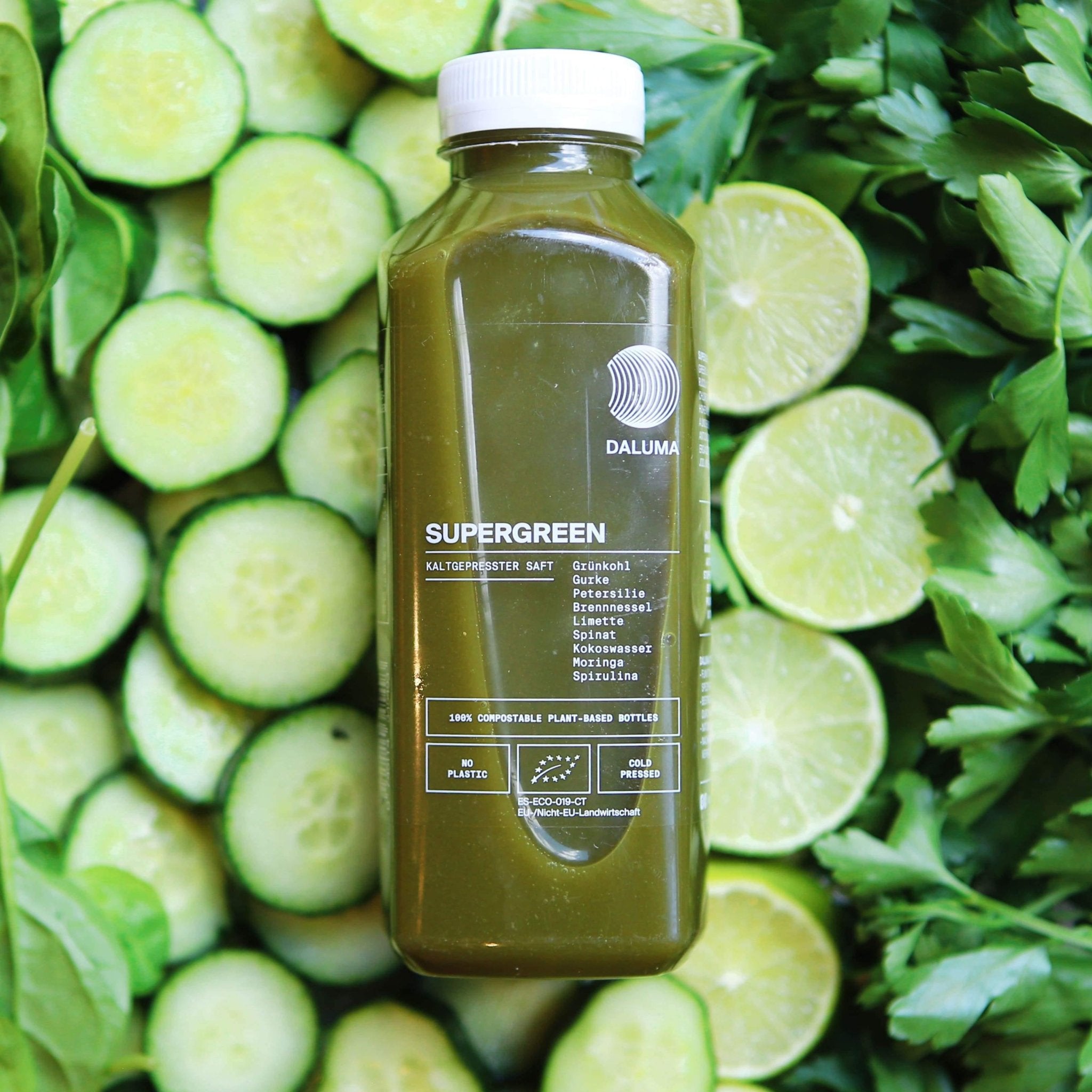 Eine Flasche des DAILY FEEL GOOD JUICE PACKAGE von DALUMA steht neben geschnittenen Gurken, Limetten und Blattgemüse. Die umweltfreundliche Verpackung hebt die kaltgepresste Säfte hervor, ideal für Ihr tägliches Abonnement.