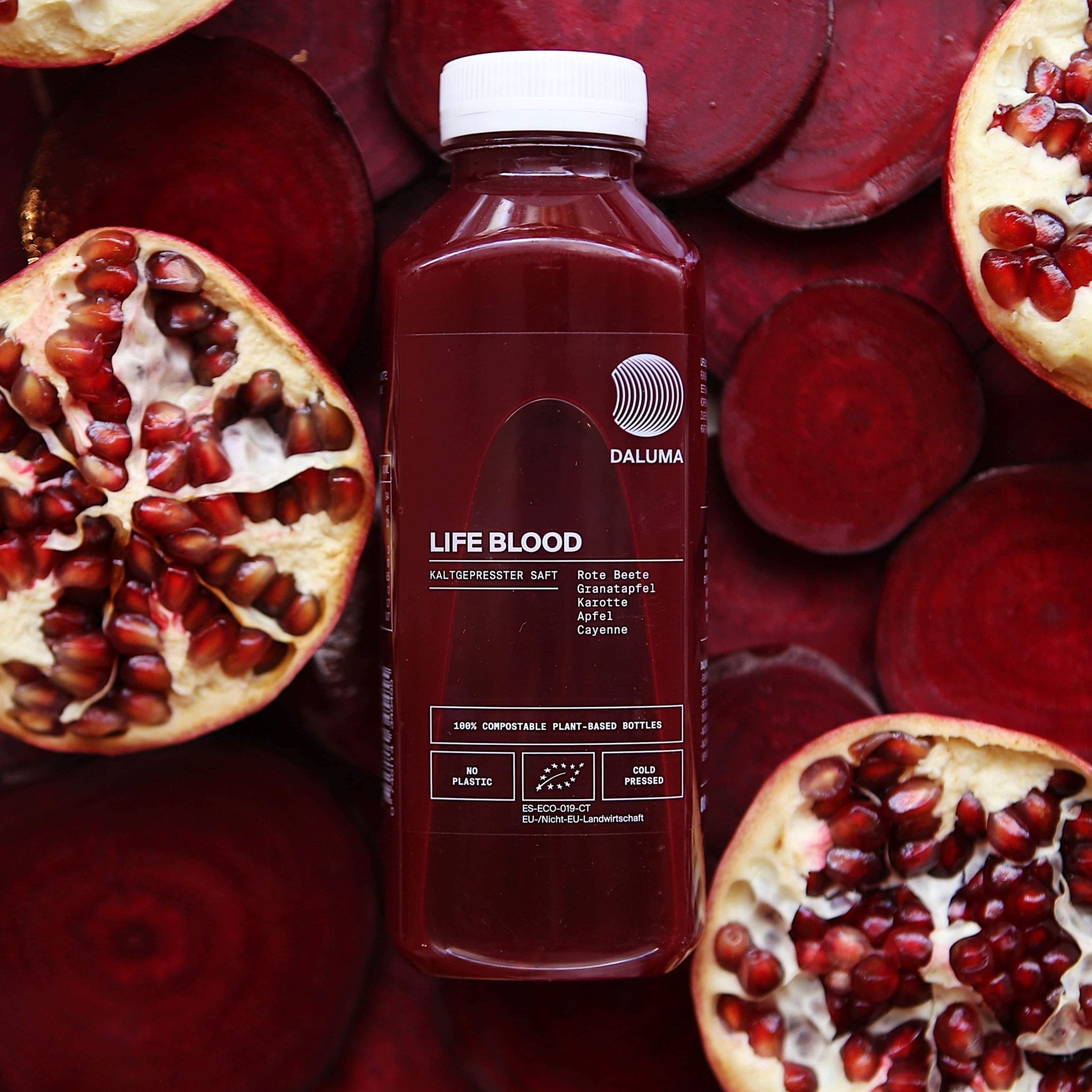 Eine Flasche DALUMA DAILY FEEL GOOD JUICE PACKAGE ist von aufgeschnittener Roter Bete und geöffneten Granatäpfeln umgeben, die die kaltgepresste Mischung aus Roter Bete, Granatapfel, Karotte, Apfel und Cayennepfeffer für echte Veggie-Saftliebhaber hervorheben.