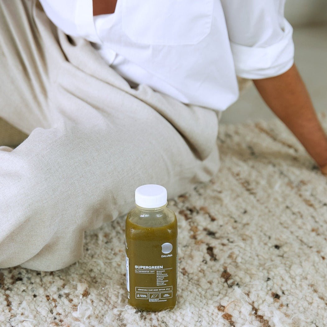 Eine Person in beiger Hose und weißem Hemd sitzt auf einem hell gemusterten Teppich, vor ihr auf dem Boden steht eine kleine Flasche DALUMA DAILY FEEL GOOD JUICE PACKAGE - perfekt für Ihr Daily Feel Good-Abo.