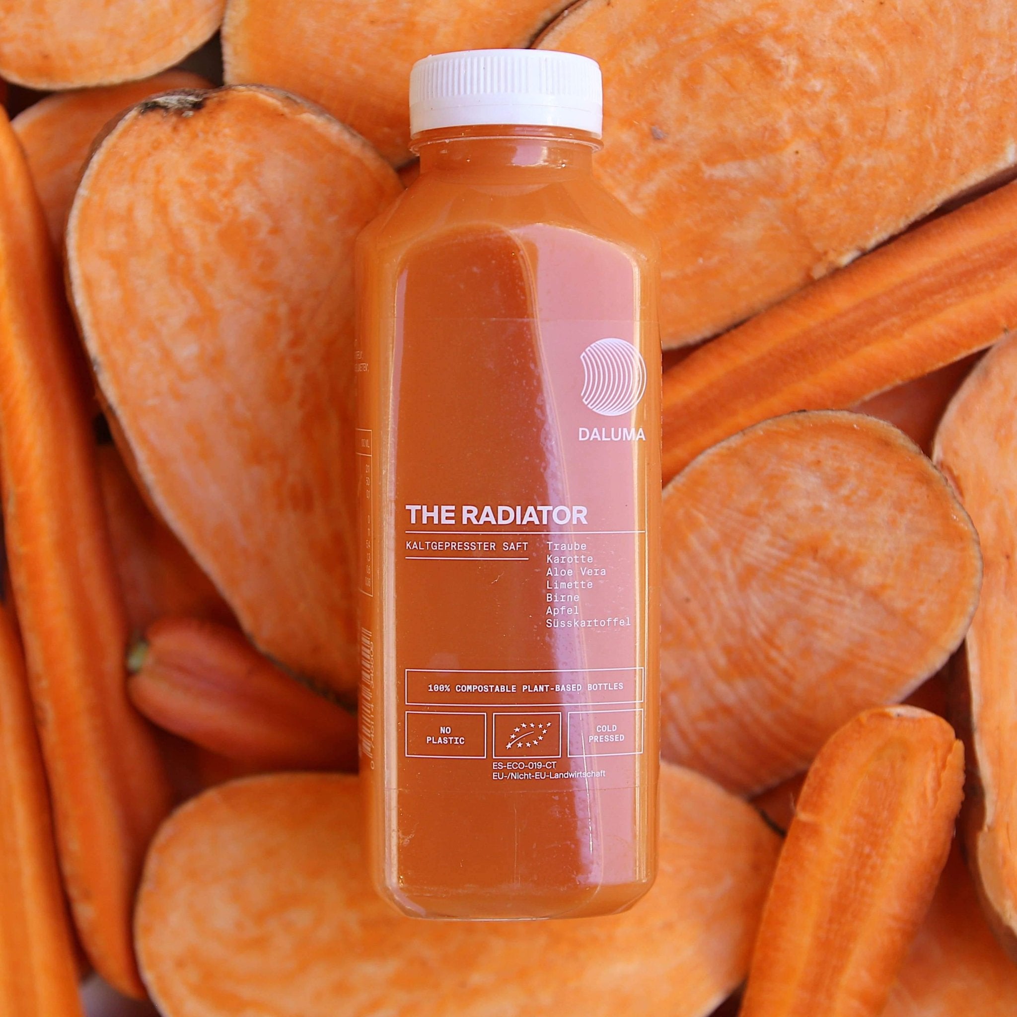 Eine Flasche des DAILY FEEL GOOD JUICE PACKAGE von DALUMA steht aufrecht auf geschnittenen Süßkartoffeln und Karotten und präsentiert ihren leuchtend orangefarbenen Farbton, die umweltfreundliche, pflanzliche Verpackung und den köstlich frischen, kalt gepressten Saft.