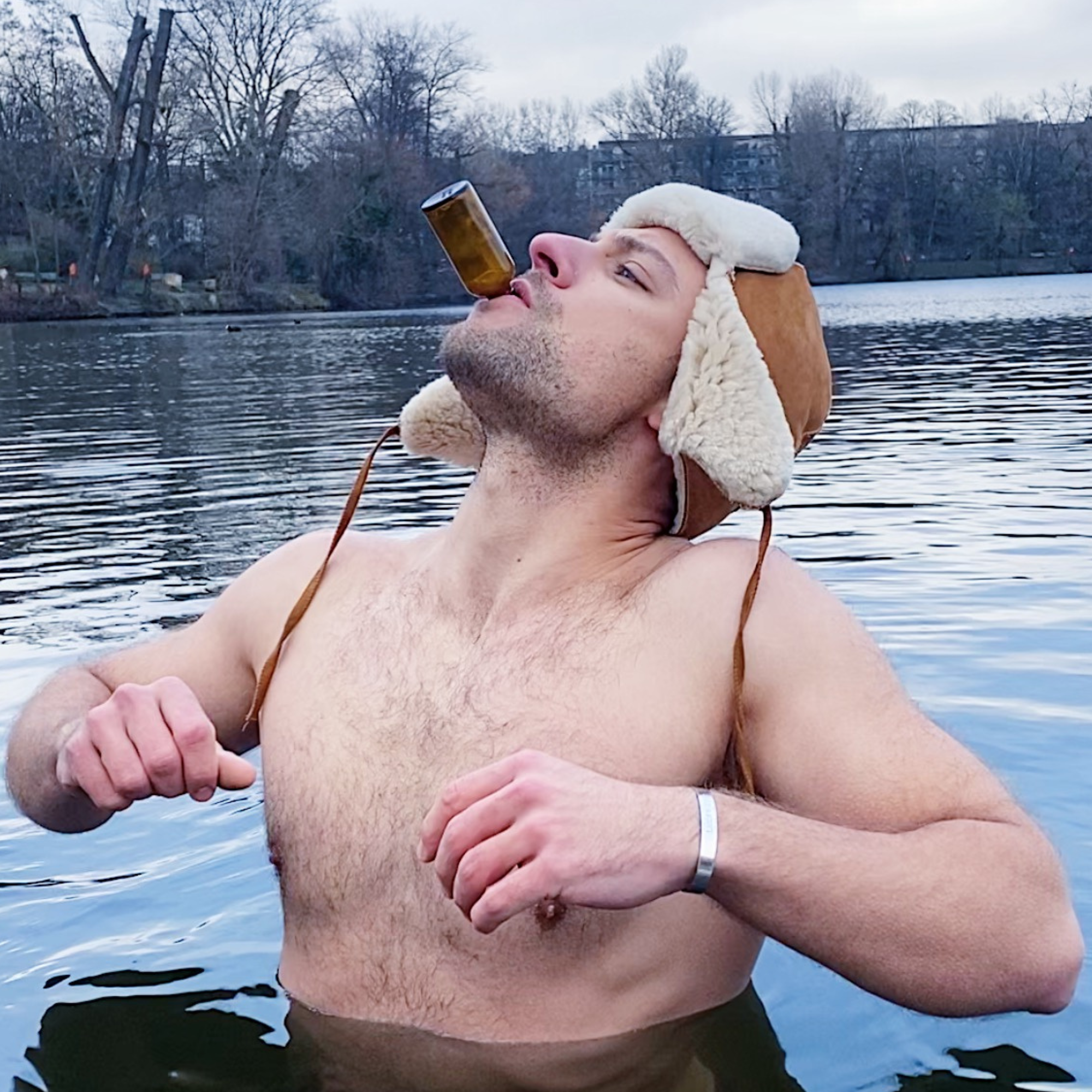 Ein Mann steht brusttief in einem See, lehnt sich zurück und hält eine kleine Flasche DALUMA BURNING MAN INGWERSHOT 24 PACK in den Mund. Im Hintergrund sind blattlose Bäume und ein wolkenverhangener Himmel zu sehen.