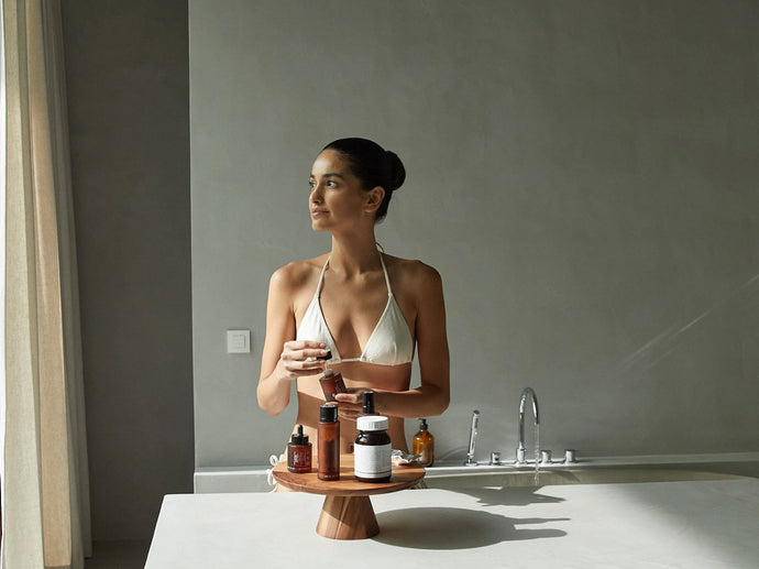 Eine Frau in einem weißen Bikinioberteil steht in einem minimalistischen Badezimmer, hält eine Tasse in der Hand und schaut zur Seite. Vor ihr auf einem Holztablett stehen mehrere Pflege- und Wellnessflaschen.