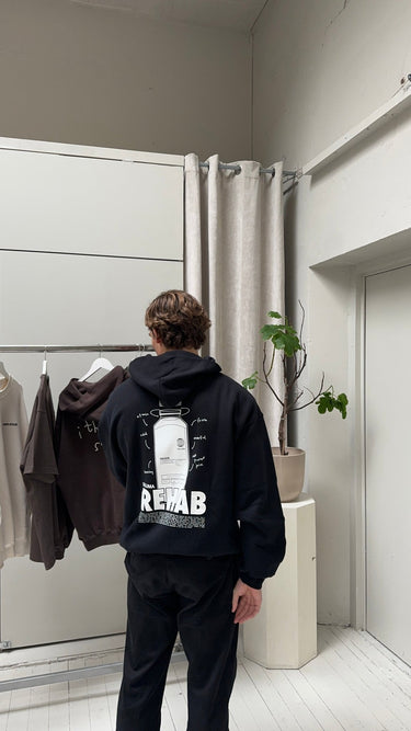 Eine Person mit kurzen braunen Haaren, die eine schwarze Hose und den DALUMA JOSEPH ATELIER X DALUMA REHAB HOODIE mit "REHAB"-Grafik auf dem Rücken trägt, steht in einem Innenraum in der Nähe eines Kleiderständers und einer Pflanze, die nachhaltige Mode präsentieren.